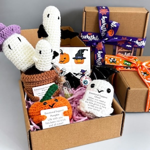 Può includere: Una confezione regalo a tema Halloween piena di oggetti all'uncinetto fatti a mano. Include un fantasma bianco, una zucca arancione e un fungo. La scatola è decorata con adesivi e un nastro viola.