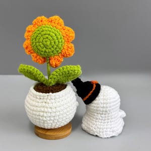 May include: A crocheted sunflower with orange petals and a green center sits in a white pot. A crocheted ghost wearing a black hat with an orange band is next to the flower. The flower pot is on a wooden base.