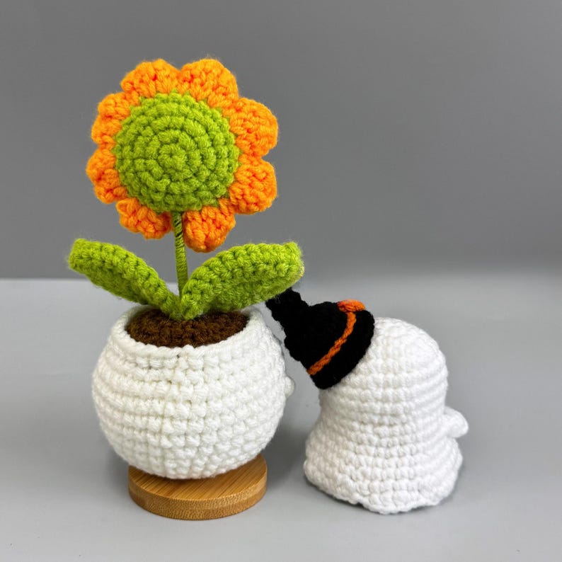 May include: A crocheted sunflower with orange petals and a green center sits in a white pot. A crocheted ghost wearing a black hat with an orange band is next to the flower. The flower pot is on a wooden base.