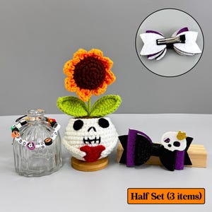 May include: A Halloween-themed set featuring a crochet sunflower in a skull pot, a clear glass bottle with a bracelet, and two hair bows. The bows are white and purple, and black and purple with a skull. The set is displayed on a neutral background.