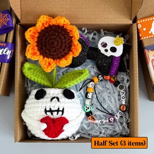 May include: A Halloween-themed gift set in a cardboard box. The set includes a crocheted sunflower, a skull-shaped hair bow, and a beaded bracelet with skull and 'BOO' beads. The box is lined with shredded paper and includes the text "Half Set (3 items)."