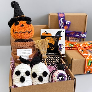 May include: A Halloween gift box featuring crocheted decorations. It contains an orange pumpkin with a black witch's hat, two white ghost figures, and a spider with a purple eye. Also included are a small broom, a card, and a music box.