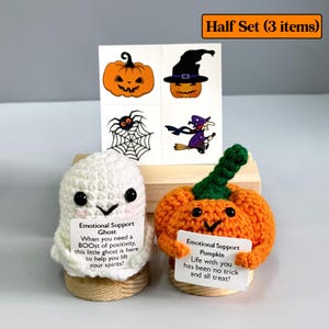 K&ouml;nnte beinhalten: Ein Halloween-Set mit einem geh&auml;kelten wei&szlig;en Geist und einem orangefarbenen K&uuml;rbis, die jeweils ein Schild mit Text halten. Enth&auml;lt tempor&auml;re Tattoos von K&uuml;rbissen, einer Spinne und einer Hexe. Das Set wird auf einem Holzst&auml;nder pr&auml;sentiert.
