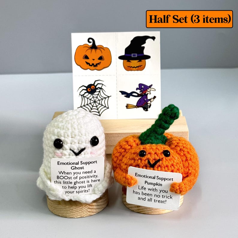 K&ouml;nnte beinhalten: Ein Halloween-Set mit einem geh&auml;kelten wei&szlig;en Geist und einem orangefarbenen K&uuml;rbis, die jeweils ein Schild mit Text halten. Enth&auml;lt tempor&auml;re Tattoos von K&uuml;rbissen, einer Spinne und einer Hexe. Das Set wird auf einem Holzst&auml;nder pr&auml;sentiert.