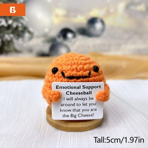 May include: Orange crocheted cheeseball holding a sign that reads "Emotional Support Cheeseball I will always be around to let you know that you are the Big Cheese!" The cheeseball is 5 cm tall.
