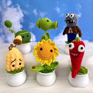 May include: A collection of crocheted plant and zombie characters from the popular video game Plants vs. Zombies. The characters include a sunflower, a pea shooter, a zombie, a chili pepper, and a corn plant. Each character is sitting in a small white pot.