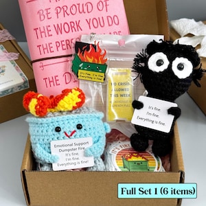 Peut inclure: Un coffret cadeau dans une boîte en carton comprend un journal rose, des jouets crochetés, des autocollants et un porte-clés. Les jouets crochetés sont un feu de poubelle bleu et une figurine noire. Le texte "It's fine, I'm fine, Everything is fine!"