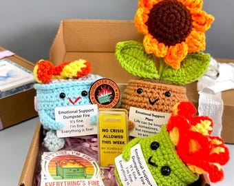 May include: A gift box filled with handmade crochet items. Includes a blue dumpster, a sunflower, and a green cactus, each with smiling faces and the text "Emotional Support Dumpster Fire." Also includes a sticker and a small sign.