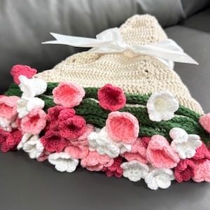 May include: A crocheted baby blanket with a floral design. The blanket is cream colored with pink and white flowers and green leaves.
