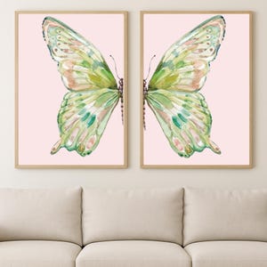 May include: Two framed butterfly art prints. Each print features a watercolor butterfly with green, pink, and peach tones against a light pink background. The frames are a light wood color, and the artwork is displayed above a beige sofa.