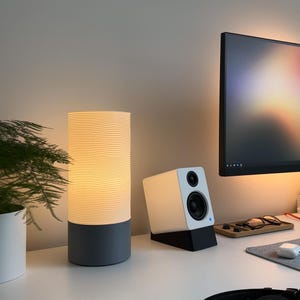 May include: A white and grey cylindrical lamp with a wavy pattern on the white shade. The lamp is on a white desk with a white speaker and a computer monitor in the background.