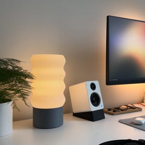 May include: A white and grey table lamp with a wavy, textured shade. The lamp is switched on and illuminating the room. A white speaker with a black base is on the table next to the lamp.