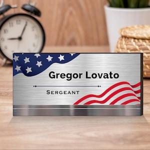 Pode incluir: Uma placa de metal prateada com acabamento escovado apresenta um design de bandeira americana e o texto "Gregor Lovato" e "SERGEANT".