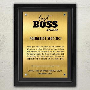 Op de afbeelding: Een ingelijste goudkleurige plaquette met de woorden "Best Boss Award" in het zwart. De plaquette is gegraveerd met een naam en een bedankbericht. De lijst is zwart en de plaquette is op een lichtgekleurde muur gemonteerd.