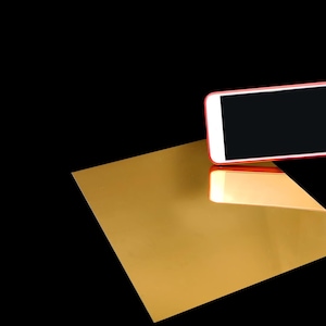 Peut inclure: Une feuille rectangulaire de mat&eacute;riau dor&eacute; r&eacute;fl&eacute;chissant avec une finition lisse semblable &agrave; un miroir. Un smartphone blanc avec un cadre rouge est positionn&eacute; au-dessus de la feuille dor&eacute;e, montrant un &eacute;cran noir et se refl&eacute;tant sur la surface.