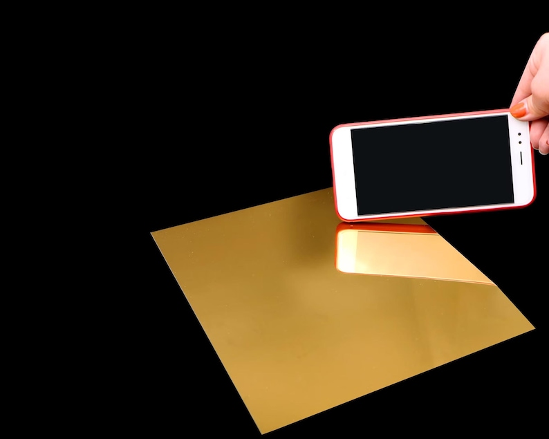 Peut inclure: Une feuille rectangulaire de mat&eacute;riau dor&eacute; r&eacute;fl&eacute;chissant avec une finition lisse semblable &agrave; un miroir. Un smartphone blanc avec un cadre rouge est positionn&eacute; au-dessus de la feuille dor&eacute;e, montrant un &eacute;cran noir et se refl&eacute;tant sur la surface.