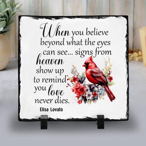 May include: A square decorative plaque with a black border and a white center. The center features a red cardinal bird perched on a floral arrangement, with the text "When you believe beyond what the eyes can see... signs from heaven show up to remind you love never dies."