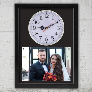 Może przedstawiać: Czarny obraz w ramie z białym zegarem i zdjęciem pary. Zegar ma czarne wskazówki i czarne cyfry rzymskie. Zdjęcie przedstawia mężczyznę i kobietę na tle rozmytego tła. Mężczyzna jest ubrany w czarny garnitur i czarną muszkę. Kobieta jest ubrana w białą sukienkę i ma długie brązowe włosy.