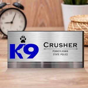 Pode incluir: Uma placa de mesa de metal prateado com o texto "K9 Crusher Pennsylvania State Police". A placa apresenta um logotipo "K9" azul com uma pegada de pata acima. A placa é retangular e tem um acabamento em metal escovado.