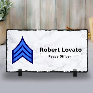Pode incluir: Placa retangular de ardósia com fundo branco efeito mármore. Apresenta uma insígnia azul em forma de galão e o texto "Robert Lovato, Peace Officer" em preto. A placa é suportada por dois suportes pretos.