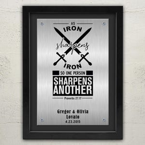 May include: A silver metal sign with black text that reads "As iron sharpens iron, so one person sharpens another - Proverbs 27:17". The sign also includes two crossed swords and the names "Gregor & Olivia Lovato" and the date "4.23.2015".
