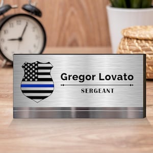 Pode incluir: Uma placa de nome de mesa prateada com borda preta. A placa de nome apresenta um distintivo de bandeira americana de linha azul fina com o texto "Gregor Lovato" e "SERGEANT" abaixo.