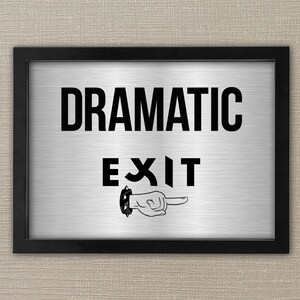 Op de afbeelding: Een ingelijst bord met de woorden "DRAMATIC EXIT" in vette zwarte letters. Het bord heeft een achtergrond van geborsteld metaal en een zwarte rand. Onder het woord "EXIT" is een hand met een wijzende vinger afgebeeld.