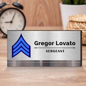 Pode incluir: Uma placa de nome de mesa prateada com um design de chevron azul e o texto "Gregor Lovato, SERGEANT".