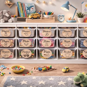 Peut inclure: Un meuble de rangement blanc avec des bacs transparents étiquetés pour les jouets, les vêtements et les fournitures d'art. Les bacs sont organisés avec des étiquettes en bois. Des jouets, un arc-en-ciel et un éléphant en peluche sont visibles.