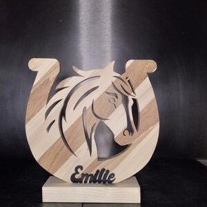 Może przedstawiać: Drewniana podkowa z wycięciem głowy konia i wygrawerowanym imieniem "Emilie" na podstawie.