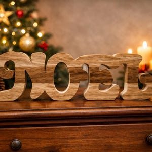 Może przedstawiać: Drewniany znak "NOEL" z dwutonowym wzorem słojów drewna, idealny do dekoracji świątecznych. Litery są wyświetlane na drewnianej powierzchni, z choinką, świecami i latarnią w tle. Świąteczny dodatek do każdej ekspozycji świątecznej.