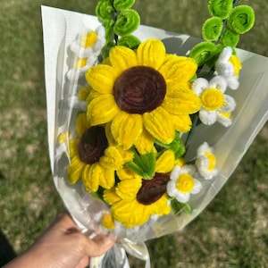May include: A bouquet of handmade sunflowers, daisies, and greenery wrapped in white paper. The sunflowers have bright yellow petals and dark brown centres. The daisies are white with yellow centres. The greenery is a bright green.