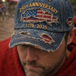 Könnte beinhalten: Verwaschene Jeans-Baseballkappe mit dem Text "250th Anniversary" und "1776-2026". Die Kappe zeigt ein zerfetztes Design der amerikanischen Flagge. Die Person trägt einen roten Kapuzenpullover.