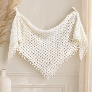 May include: A triangular off-white crochet shawl featuring an intricate openwork pattern and a delicate border of small, dangling pom-poms along the bottom edges. The shawl is suspended by two knotted corners, showcasing its lightweight, airy texture.