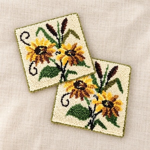 Könnte beinhalten: Zwei quadratische Punch-Needle-Untersetzer mit cremefarbenem Hintergrund und Sonnenblumenmotiv. Jeder Untersetzer zeigt zwei gelbe Sonnenblumen mit dunkelbraunen Mitten, grünen Blättern und dunkelbraunen Stielen, eingefasst in einen grünen Rand.