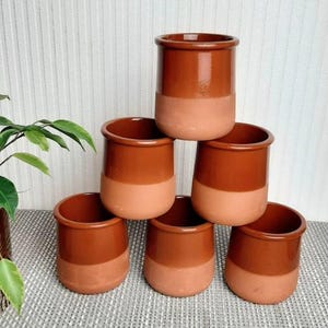 May include: A stack of six terracotta-colored ceramic pots. Each pot has a cylindrical shape with a rounded rim and a two-tone design, featuring a darker brown upper half and a lighter, natural clay-colored lower half. The pots are arranged in a pyramid shape.
