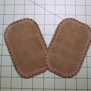 May include: Two brown suede patches with a stitched edge. The patches are oval shaped and have a smooth surface.