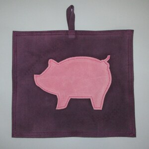 May include: A purple suede pot holder with a pink pig silhouette sewn on. The pot holder has a loop for hanging.