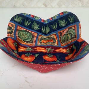 Puede incluir: Un bol de tela rojo y azul con un forro de lunares blancos. El bol presenta un estampado vintage de verduras, incluyendo tomates, col y judías verdes. El texto en el bol dice "Quality Grown Cabbage", "Tomatoes California" y "Butterflied Stringless Beans".