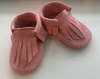 May include: A pair of pink suede baby moccasins with fringe. The moccasins have a pink stitching detail.