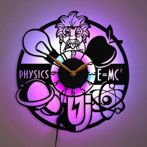 May include: A black and white clock with a science theme. The clock features a cartoon image of Albert Einstein, a light bulb, an apple, a planet, a magnet, and an atom. The clock face is black with white numbers and a gold-coloured hour and minute hand. The clock has a white background with the words "PHYSICS" and "E=MC^2" printed on it.