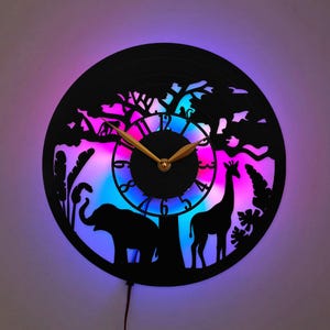 Op de afbeelding: Een ronde wandklok met een zwarte silhouet-uitsnede van een olifant, een giraf en bomen tegen een levendige, gloeiende LED-achtergrond met paars en blauw licht. De wijzerplaat heeft zwarte cijfers en goudkleurige wijzers.