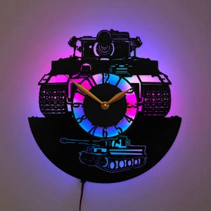 Puede incluir: Un reloj de pared circular hecho de un disco de vinilo negro, con una silueta de tanque cortada con láser. La esfera tiene manecillas doradas y está iluminada por una luz LED multicolor vibrante que brilla en tonos azules y rosados.