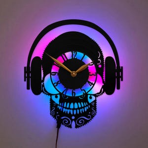 Può includere: Un orologio da parete realizzato con un disco in vinile nero, a forma di teschio che indossa cuffie. Il quadrante presenta lancette e numeri color oro, illuminati da dietro da una vivace luce LED di colore viola e blu.