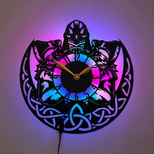May include: A circular wall clock featuring a black cutout design of a Viking warrior holding two axes. The clock face has gold-colored hands and is illuminated by a vibrant purple and blue LED light that highlights the intricate Celtic knotwork.