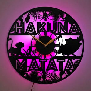 Puede incluir: Un reloj de pared negro y dorado con una silueta de Simba y Pumbaa de la película El Rey León. La esfera del reloj tiene números romanos y las palabras "Hakuna Matata" se muestran arriba y abajo de la esfera. El reloj está iluminado con luces LED rosas.