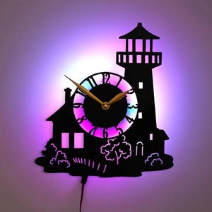 Puede incluir: Un reloj de pared negro con temática de faro con un fondo iluminado colorido. El reloj presenta un diseño de silueta con una casa, árboles y un faro. Las manecillas del reloj son doradas y el reloj funciona con un cable rojo.