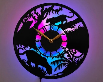 May include: A circular wall clock crafted from a black vinyl record featuring a laser-cut silhouette of panthers in a jungle setting. The clock face has gold-colored hands and is illuminated from behind by a vibrant purple and blue LED light.
