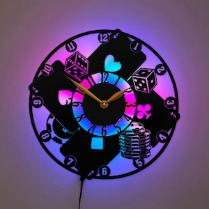 May include: A circular wall clock crafted from a vinyl record featuring a casino theme with cutouts of playing cards, dice, and poker chips. The clock face displays numbers one through twelve and is illuminated by a vibrant, multi-colored LED light.