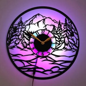 May include: A black circular wall clock featuring a mountain scene with a colourful LED light. The clock has a black background with a white mountain range, trees, and a river. The clock face is black with white numbers and gold hands. The LED light is multi-coloured and creates a colourful glow around the clock.